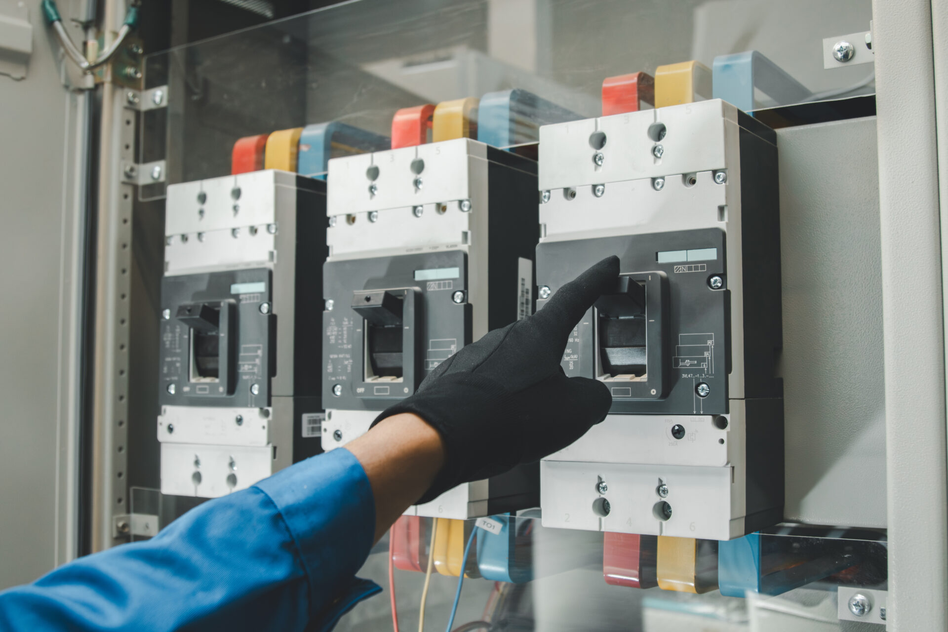 A person wearing a black glove is operating a switch on an industrial electrical circuit breaker panel with multiple switches and colored wiring, showcasing expert switchgear installation & repair.