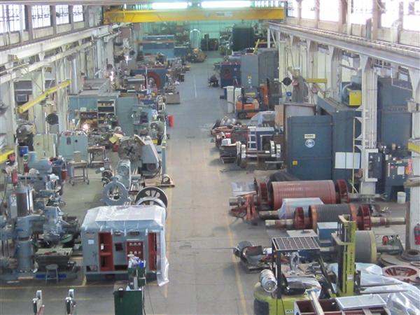 about-spot An industrial workshop with various machinery and equipment arranged along the floor. Several large components are scattered throughout the space, with some wrapped in plastic. Overhead cranes and storage areas are visible in the background.