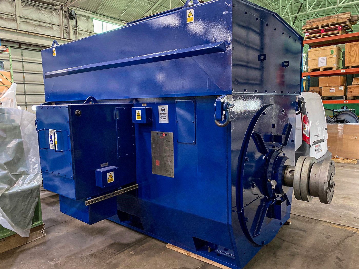 equipment-new-motor Large industrial blue electric motor with a metal shaft, sitting on a warehouse floor. Surrounding the motor are various storage crates and equipment within a spacious industrial facility.