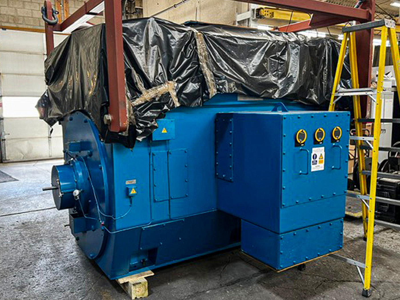 A large blue industrial machine, partially covered with black plastic, stands inside a workshop. A yellow ladder is positioned next to it, and the background shows a garage door and concrete walls.